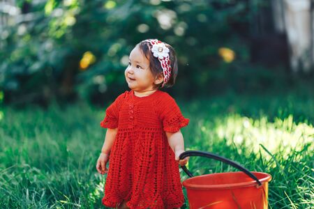 Adorable happy toddler girl with knitten flower crown wearing a red dress enjoying picnic in a beautiful blooming fruit garden.の写真素材