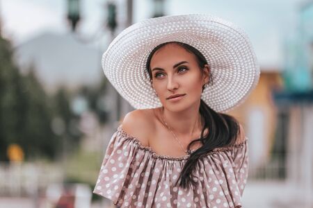 Young beautiful woman with long hair in white hat and begie dress walking in a city.の写真素材