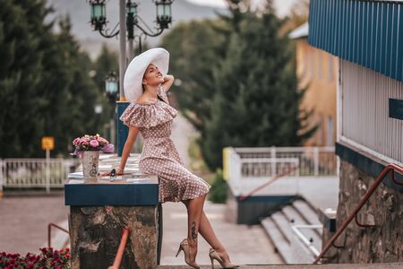 Young beautiful woman with long hair in white hat and begie dress walking in a city.の写真素材