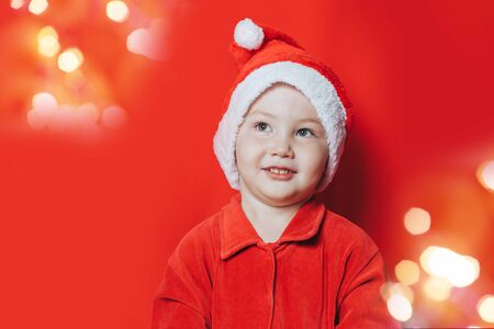 Small smiling boy wearing in Santa hat and suit looking at camera and waiting christmas on red background.の写真素材