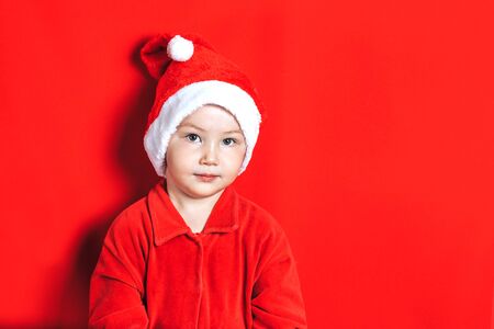 Small smiling boy wearing in Santa hat and suit looking at camera and waiting christmas on red background.の写真素材