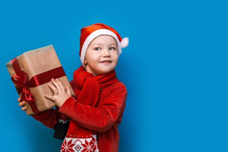 Small blonde boy in Santa suit holding christmas gift with red ribbon on blue background.の写真素材