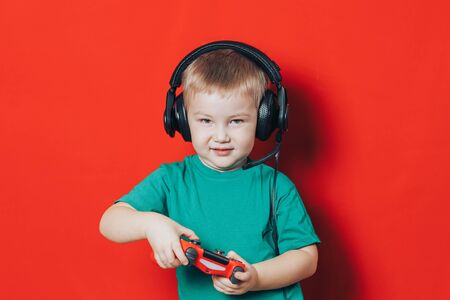 Little boy playing with red joystick have angry emotions on his face, in headphones on red...