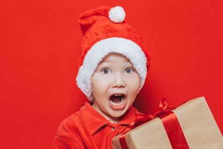 Small blonde boy in Santa suit holding christmas gift with red ribbon on red background. Very surprised kid because of holiday or gift.の写真素材