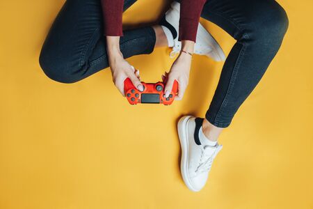 Young thin woman playing with red gamepad, sitting on yellow background. flat lay.