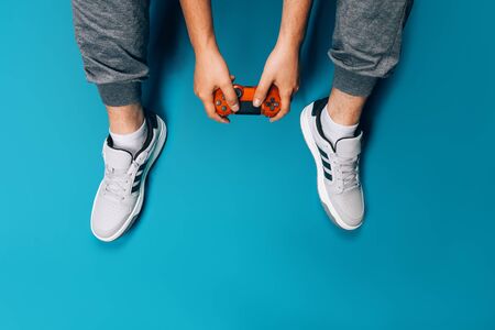 A young guy plays video games in his hands holding a red gamepad on a blue background, sitting in...