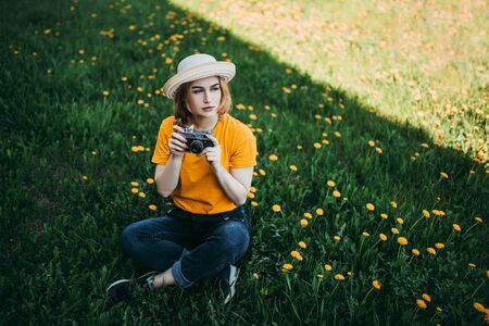 Young beautiful girl in an orange T-shirt holding retro camera in her hands lying on the lawn where dandelions grow, flowering dandelionsの写真素材