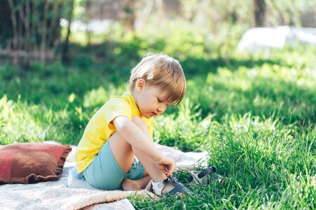 Beautiful little boy takes off shoes while sitting on a lawn in the open air on a sunny dayの写真素材