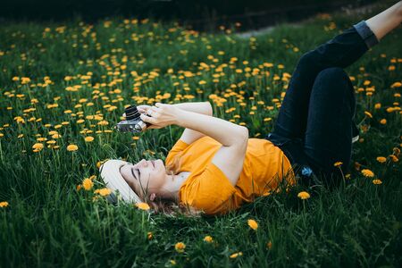 Young beautiful girl in an orange T-shirt holding retro camera in her hands lying on the lawn where dandelions grow, flowering dandelionsの写真素材