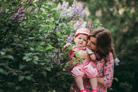 Young beautiful mother with small child in pink clothes on the background of beautiful blooming lilac in the openの写真素材