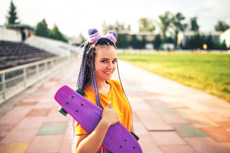 Young beautiful girl holding a skateboard in a hand in a yellow t-shirt in the open air at the stadium.の写真素材