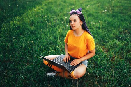 Young beautiful girl holding a laptop in her hands in a yellow T-shirt sitting on a lawn in the open airの写真素材