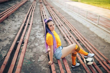 Beautiful young girl with long hair and a soccer ball sitting in a stadium, unusual hairstyle.の写真素材