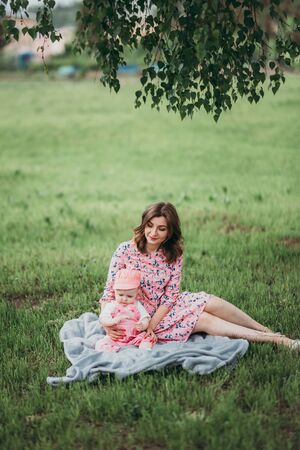 Young beautiful mother with small child have rest in the park on the lawn on warm day in pink clothes under the open sky.の写真素材