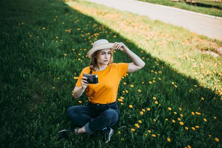 Young beautiful girl in an orange T-shirt holding retro camera in her hands lying on the lawn where dandelions grow, flowering dandelions.の写真素材