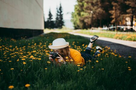 Young beautiful girl in an orange T-shirt holding retro camera in her hands lying on the lawn where dandelions grow, flowering dandelions.の写真素材