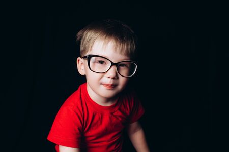 Pretty blonde boy sitting on black background in glasses, Myopia. Kids visionの写真素材
