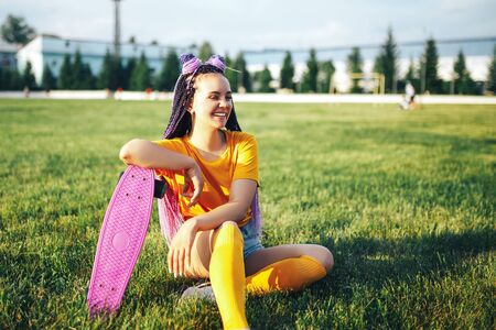 Young beautiful girl sitting on a lawn holding a skateboard in her hand in a yellow T-shirt and yellow socks in the openの写真素材
