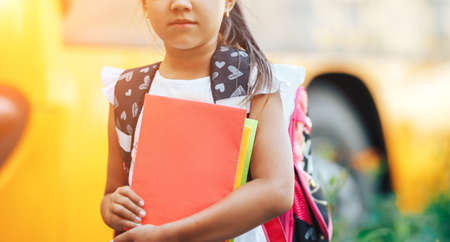 A schoolgirl with a backpack holding a book in her hands on the background of a school bus, back to school.の写真素材