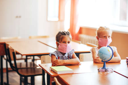 Two little schoolgirls sit at a desk in a school class in medical masks and carefully listen to the teacherの写真素材