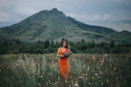 A girl in an orange dress with a bouquet of flowers walking along the field against the background of grass and mountains.の写真素材