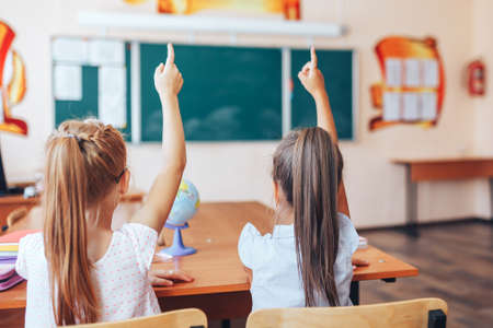 Two little schoolgirls sit at a desk in a school class and raise their hands.の写真素材