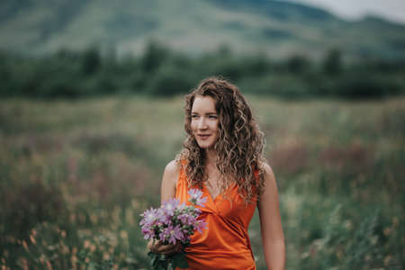 A girl in an orange dress with a bouquet of flowers walking along the field against the background of grass and mountains.の写真素材