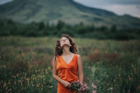 A girl in an orange dress with a bouquet of flowers walking along the field against the background of grass and mountains.の写真素材