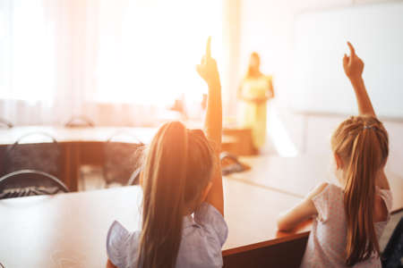 Two little schoolgirls sit at a desk in a school class and raise their hands.の写真素材