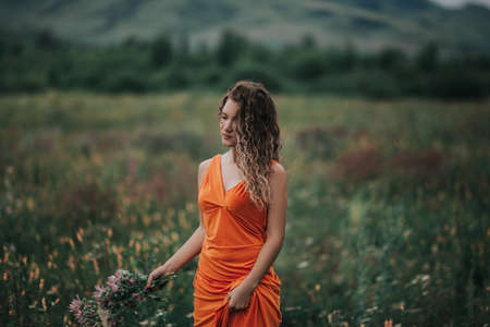 A girl in an orange dress with a bouquet of flowers walking along the field against the background of grass and mountains.の写真素材