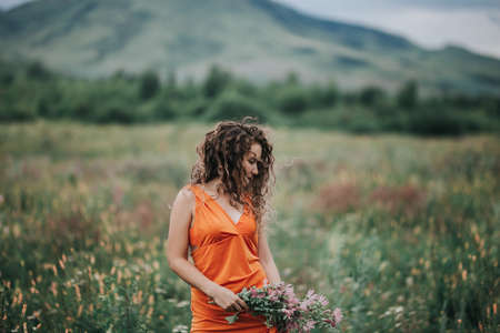 A girl in an orange dress with a bouquet of flowers walking along the field against the background of grass and mountains.の写真素材
