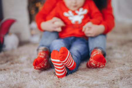 Mom holding her child by the hands sitting on a soft carpet in red sweaters and red socks, Merry...