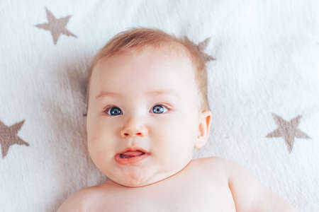 A baby grimaces at the camera, a newborn baby with blue eyes and blond hair lying on a light blanket with brown starsの写真素材