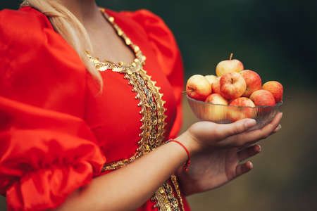 A young girl dressed in national Russian clothes of red color outdoors holding a plate with apples in her hand. Art grain and noise on photoの写真素材