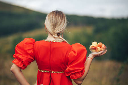A young girl dressed in national Russian clothes of red color outdoors holding a plate with apples...