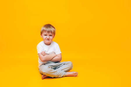 Crying little boy rubs his eyes with his hands, a boy in a white t-shirt on a yellow background.