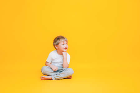 Pensive little boy in white t-shirt sitting on yellow background.の写真素材