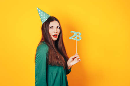 Woman with surprised face in one hand holds the face and the other holds the number 28 in green dress on yellow background.の写真素材