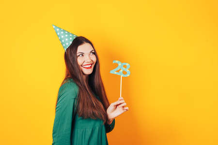 Beautiful woman celebrates her birthday on a yellow background in green dress and on her head festive cap holds the number 28 in her hand.の写真素材