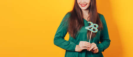 Beautiful woman celebrates her birthday on a yellow background in green dress and on her head festive cap holds the number 28 in her hand.の写真素材