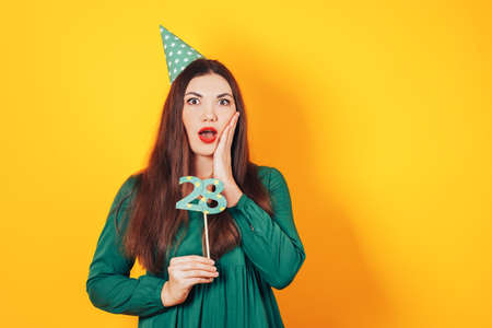 Woman with surprised face in one hand holds the face and the other holds the number 28 in green dress on yellow background.の写真素材