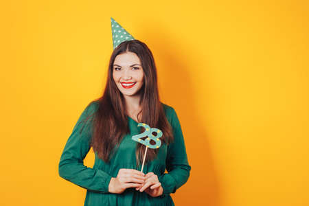Beautiful woman celebrates her birthday on a yellow background in green dress and on her head festive cap holds the number 28 in her hand.の写真素材