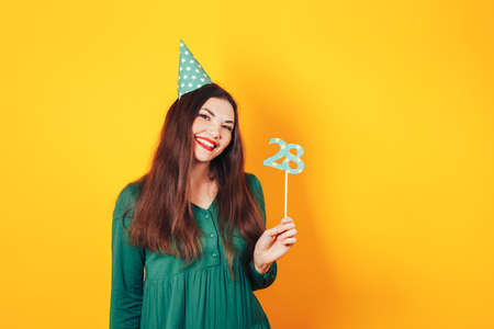 Beautiful woman celebrates her birthday on a yellow background in green dress and on her head festive cap holds the number 28 in her hand.の写真素材