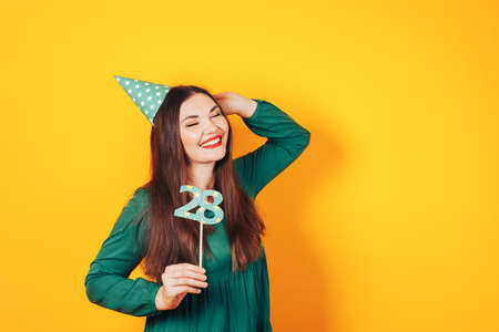 Beautiful woman celebrates her birthday on a yellow background in green dress and on her head festive cap holds the number 28 in her hand.の写真素材
