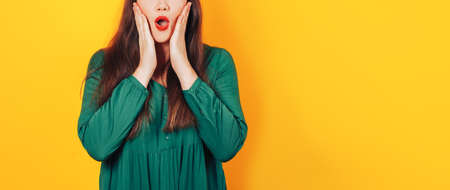 Woman with a surprised face holding her face with her hands in a green dress on a yellow background.の写真素材