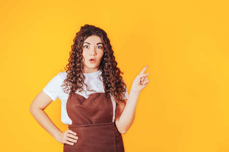 Young woman dressed in barista uniform pointing finger to the side on yellow background.の写真素材