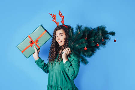 Young woman holding a big gift and a Christmas tree in her hands, she has deer horns on her head on a blue backgroundの写真素材