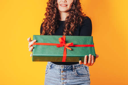 Beautiful woman in black jacket and red hat on her head on yellow background holding large box with gift in her hands.の写真素材