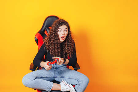 Woman sitting behind gaming chair in her hands holding red gamepad on yellow background.