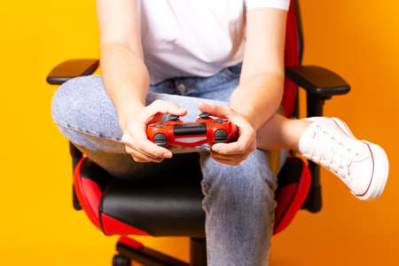Woman sitting behind gaming chair in her hands holding red gamepad on yellow background.